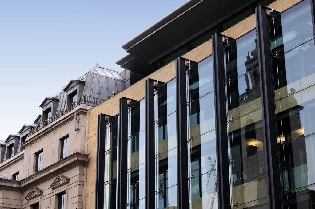 Modern glass building with reflective windows is adjacent to an older, beige stone building with a mansard roof and decorative architectural details under a clear sky.