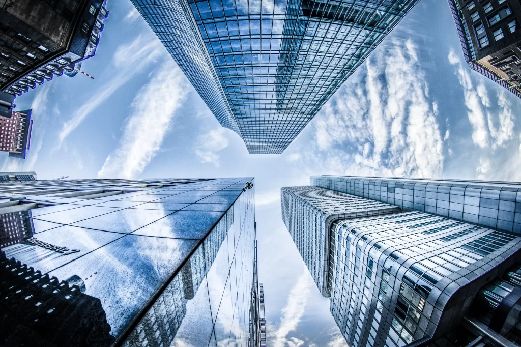 Skyscrapers viewed from below with glass facades reflecting clouds and a blue sky, creating a dramatic urban perspective.